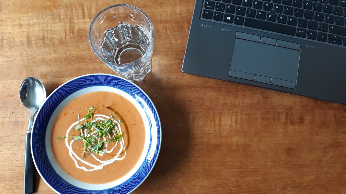 Skål med soppa framdukad framför en laptop på ett träbord.